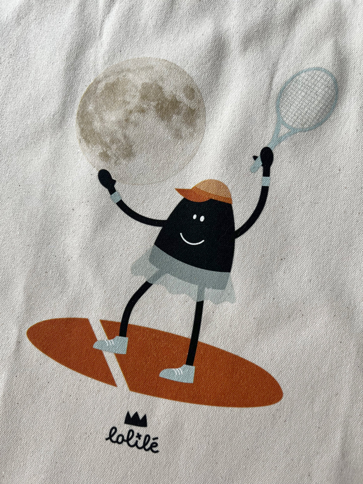 Tote bag « Jeu, set et lune » de Lolilé, représentant un petit personnage noir en tenue de tennis tenant une raquette et jouant avec une grande lune sur fond quadrillé bleu clair.