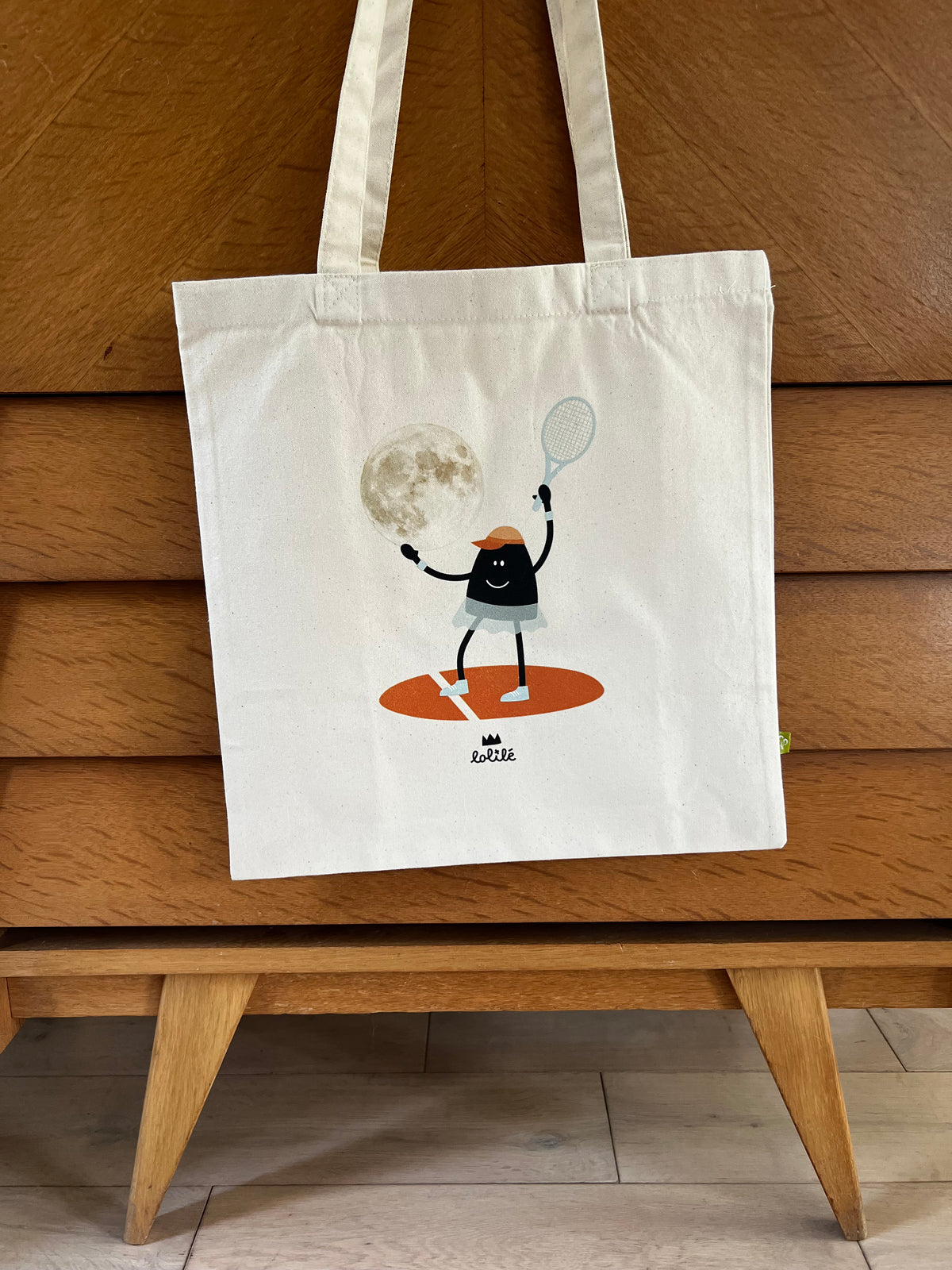 Tote bag « Jeu, set et lune » de Lolilé, représentant un petit personnage noir en tenue de tennis tenant une raquette et jouant avec une grande lune sur fond quadrillé bleu clair.
